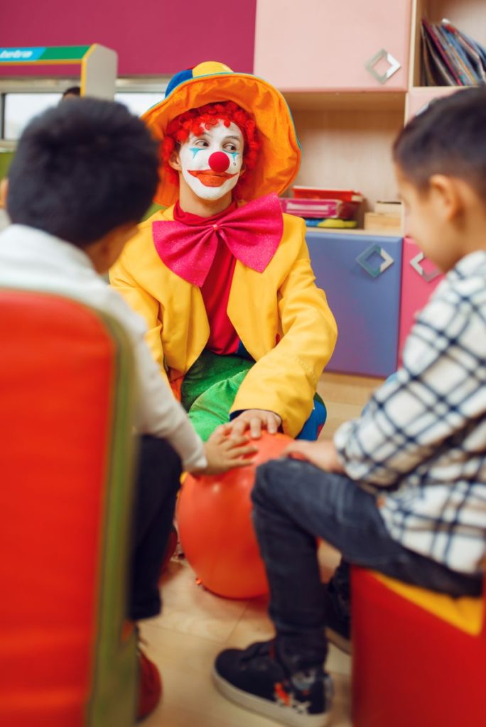 Funny clown play with cheerful children together. Birthday party celebrating in playroom, baby holiday in playground. Childhood happiness