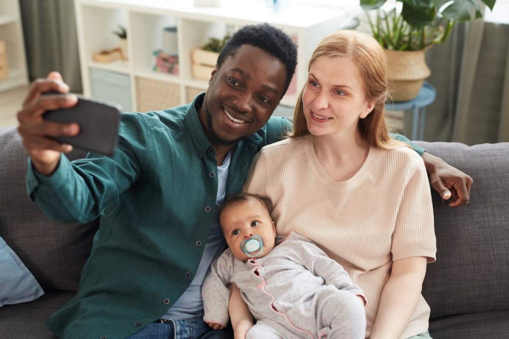 High angle portrait of young interracial family taking selfie photo with cute mixed-race baby while sitting on couch in home interior